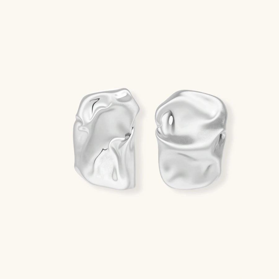 Pair of sculptural silver ear cuffs with organic fluid shapes and polished finish against cream background.